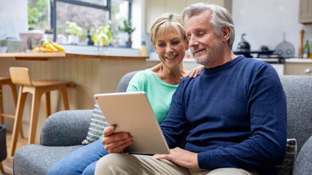 A senior couple sit on a couch at home looking at a laptop.