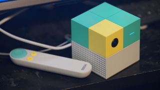 The Nex Playground console on a shelf, showcasing its cubic build with in-built camera and LED light. The included Nex remote sits next to it, featuring a dial and a couple of buttons.