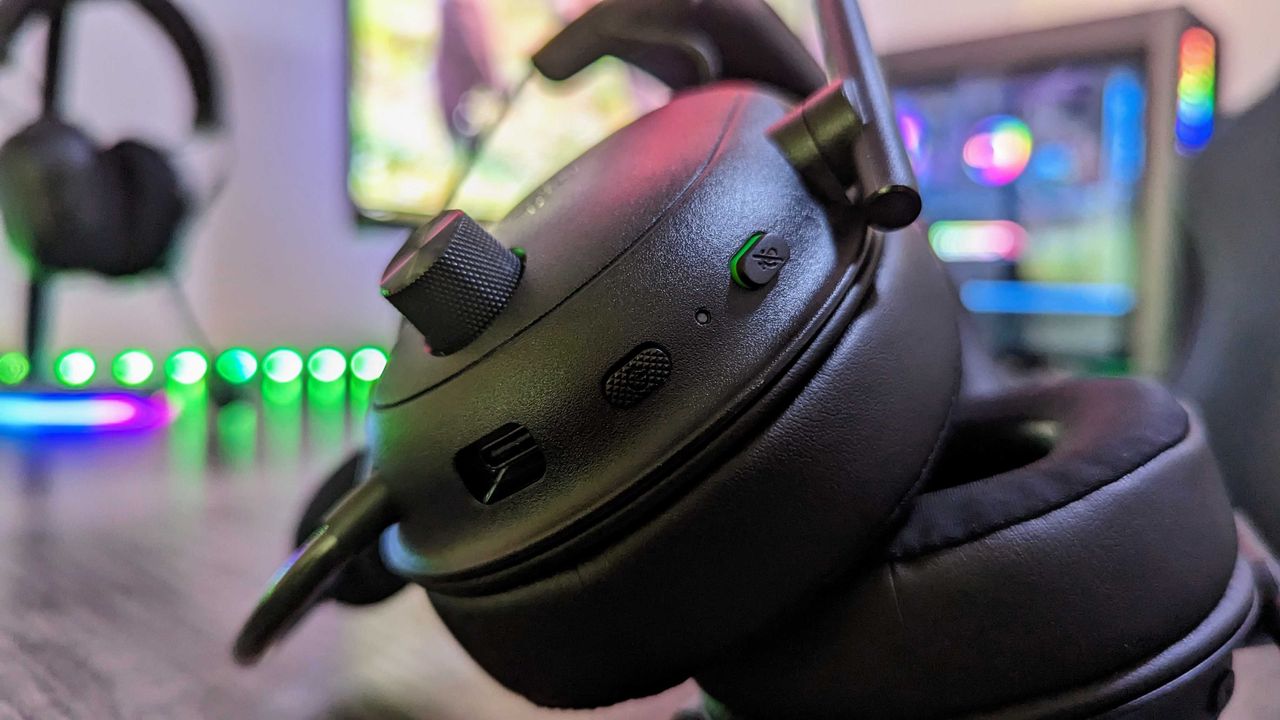 Close-up of black over-ear headphones with control buttons and volume knob, set against a blurred, colorful gaming setup, conveying a tech-savvy vibe.