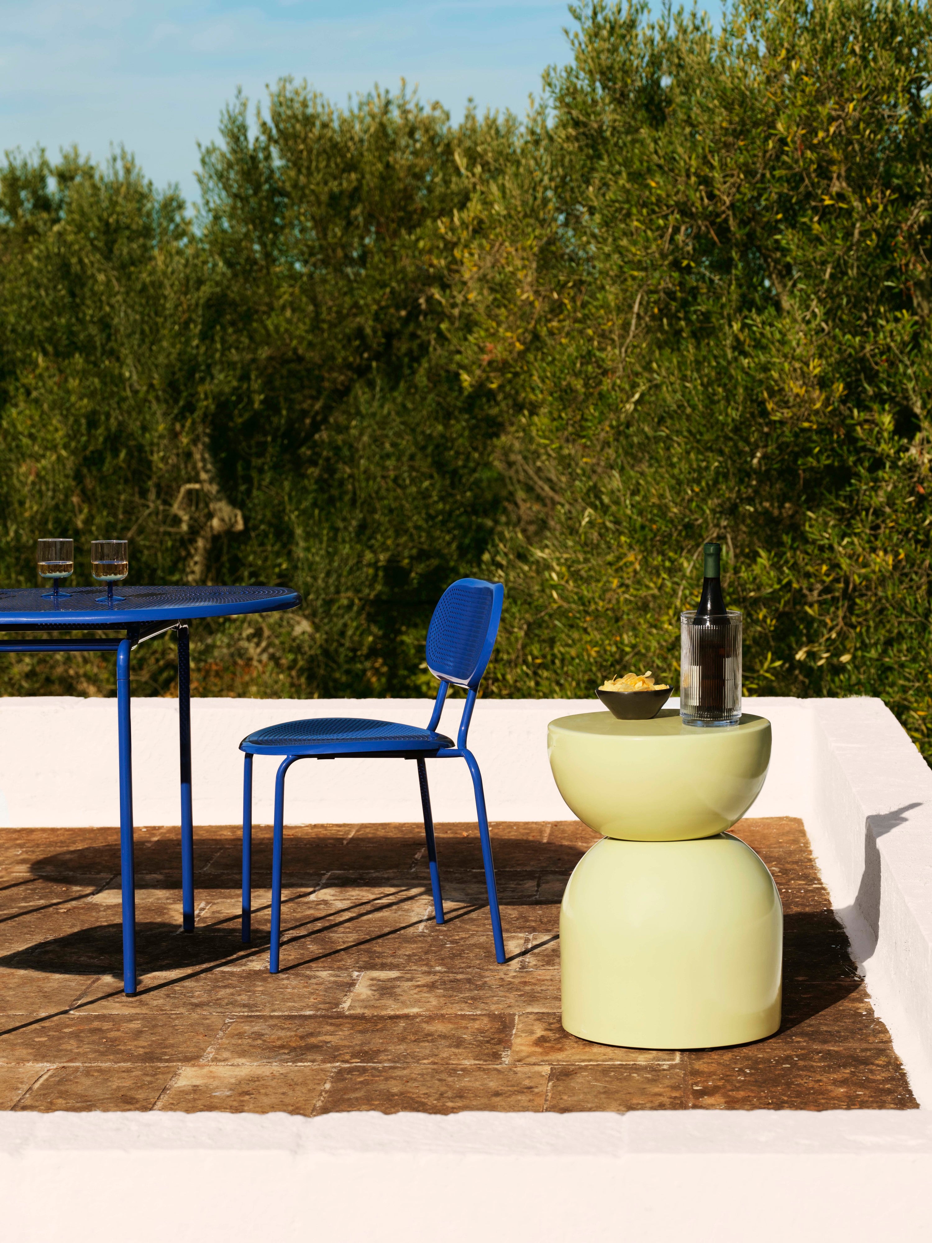Image of an outdoor patio by the sea with a royal blue bistro set and a light yellow side table that has a wine bottle on it.