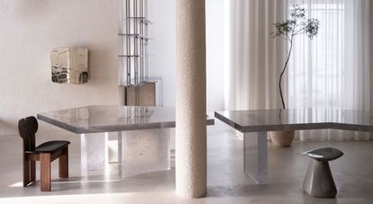 A showroom with a central cement column, decorated with geometrical glass tables, sculptural chairs in wood and leather and polished stone, reflective artworks, and a plant potted in ceramic vase.