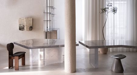 A showroom with a central cement column, decorated with geometrical glass tables, sculptural chairs in wood and leather and polished stone, reflective artworks, and a plant potted in ceramic vase.