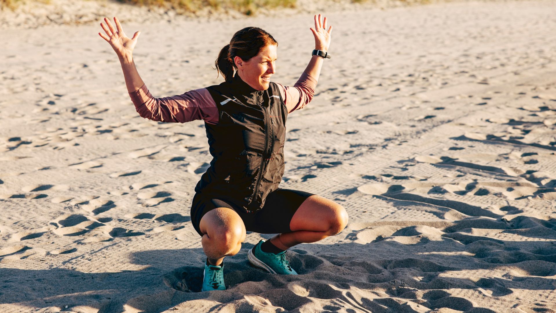Woman at the bottom of a squat with hands in the air on the beach