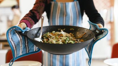 A person holding a wok full of healthy stir fry vegetables with oven gloves