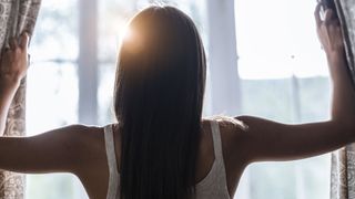 A woman with long brown hair opens the curtains in her bedroom to let in natural light straight after waking up