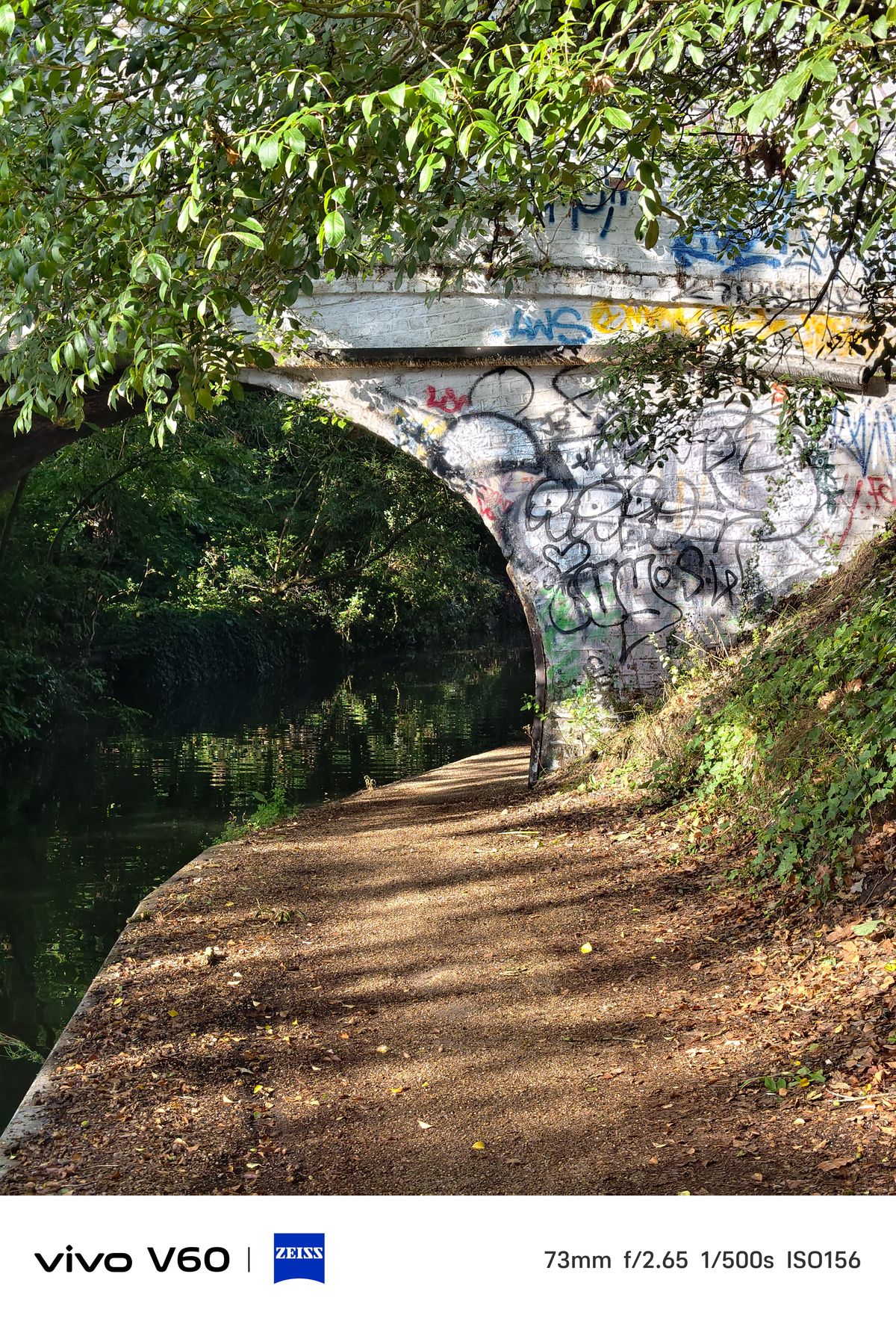 A canal path leading under a bridge covered in graffiti