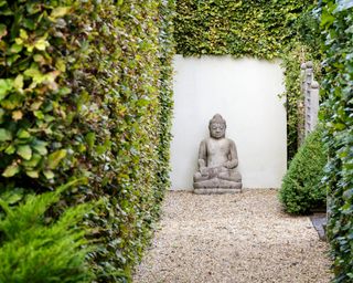 A Buddha statue at the end of a long gravel path lined with hedges.