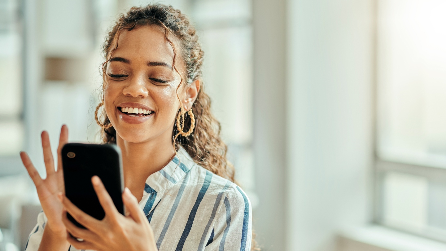 Woman with brown curly hair holding phone and smiling at it