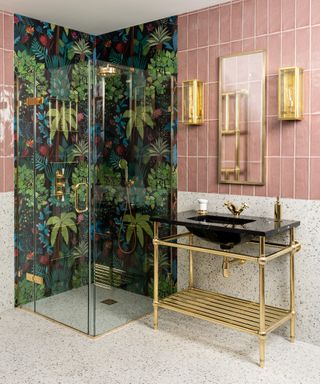 An example of bathroom shelf ideas showing a pink-tiled bathroom with walk-in shower clad with tropical patterned wallpaper, and an open gold vanity unit