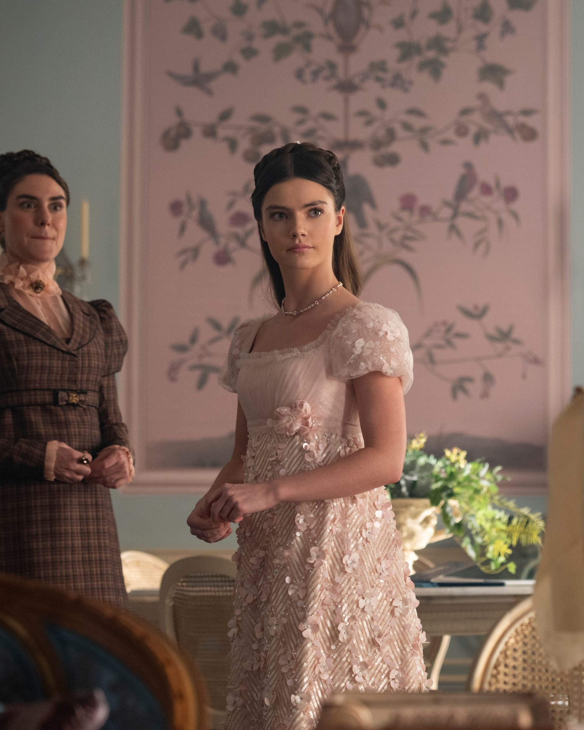 Production still from Netflix's Bridgerton series showing a girl in a light pink gown standing in a blue room that has a pink floral panel of wallpaper on the wall.