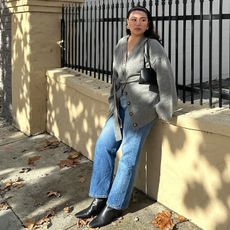 Marina wears blue jeans, gray sweater with belt, and black square toe boots and a black handbag while posing for the camera in her outfit. 