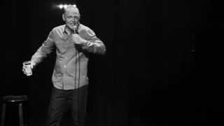 Blck and white shot of Burr on stage holding a glass of water in his hand.