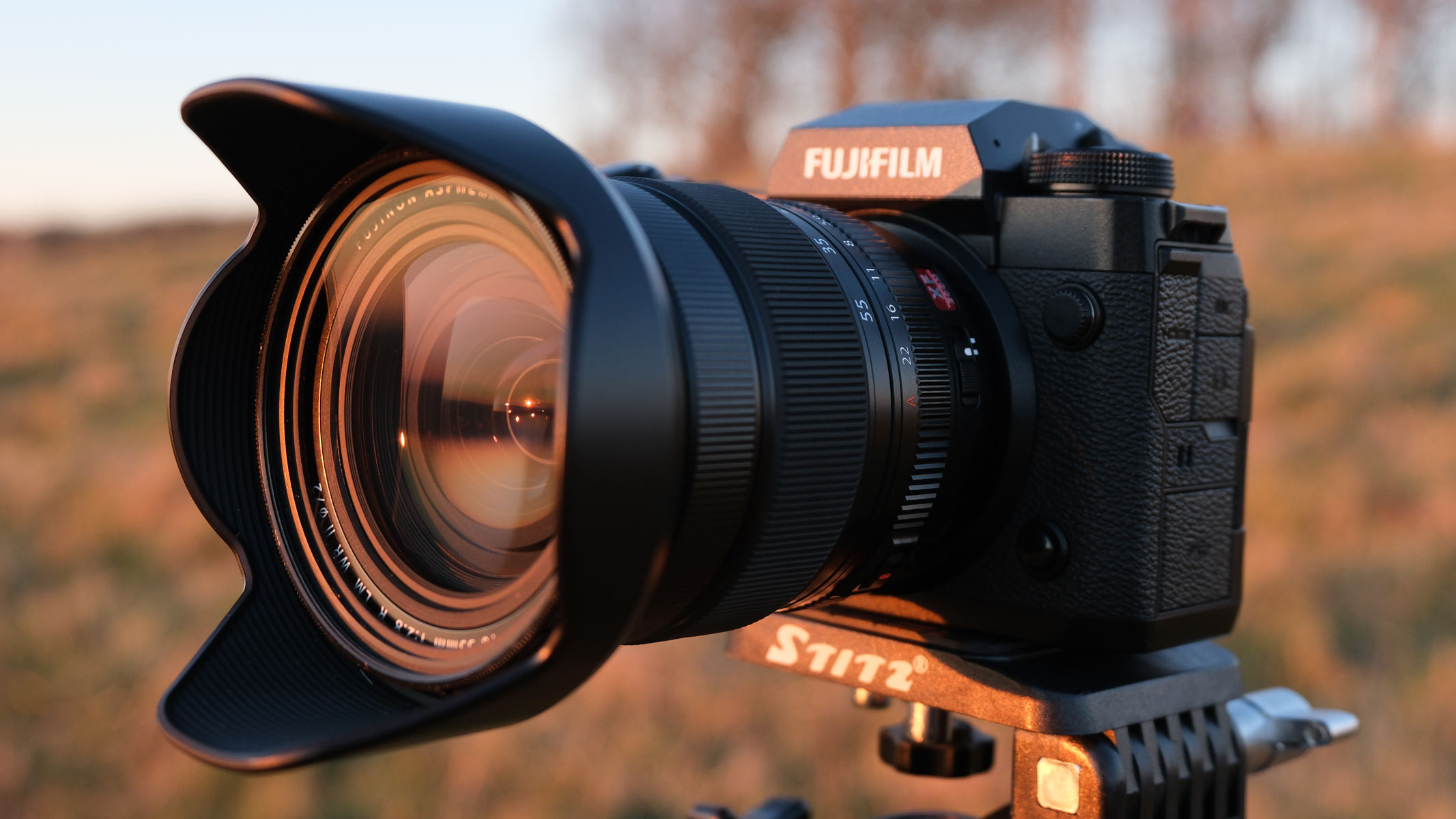 A side-on view of the Fujifilm X-H2 with the Fujifilm XF 16-55mm f/2.8 LM WR II lens attached and a field in the background