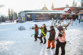 Four people walking through a ski resort carrying skis over their shoulders