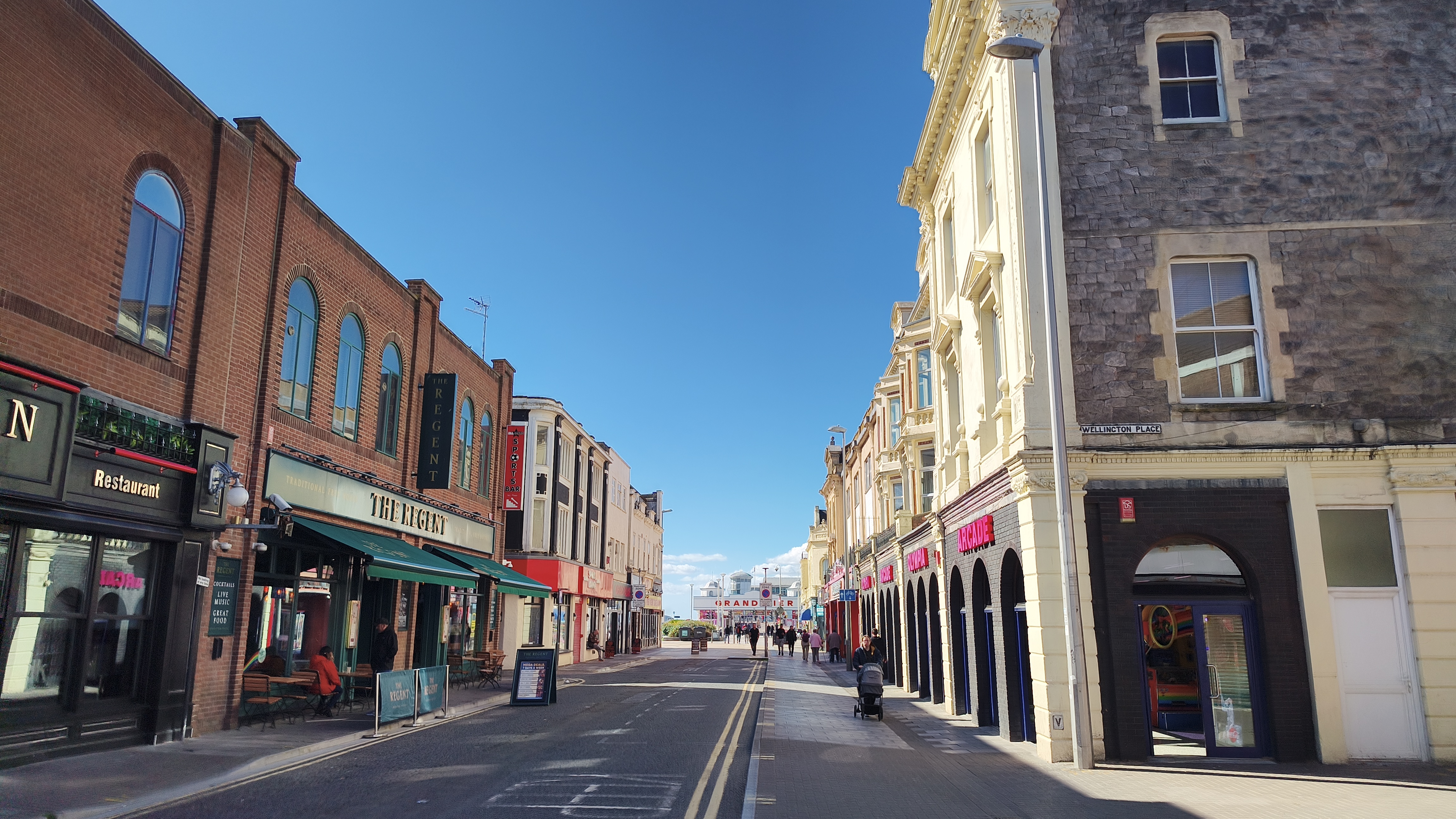 This photo shows a sunny, street-level view of shops and restaurants with people strolling along the sidewalk.