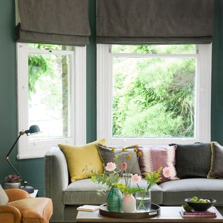 cosy living room with green painted d walls and a sage green sofa filled with pink and yellow cushions a heavy lined roman blind in the window and flowers in a vase on the table