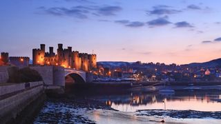 A classic dusk shot of Conwy Castle in Wales, with bridge leading in and town lights glowing in the background