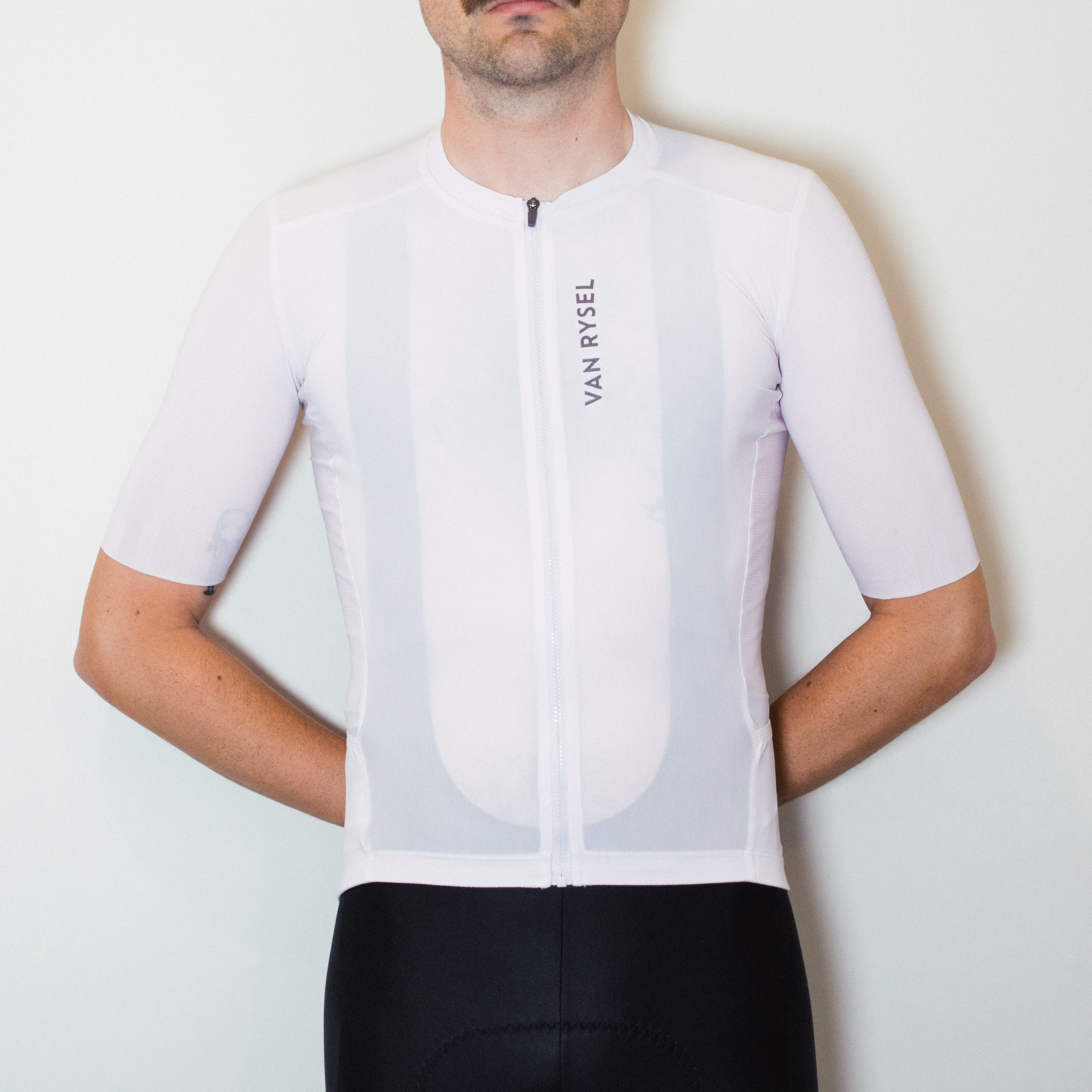 A white man stands against a white wall wearing black cycling shorts and a tight pale pink jersey