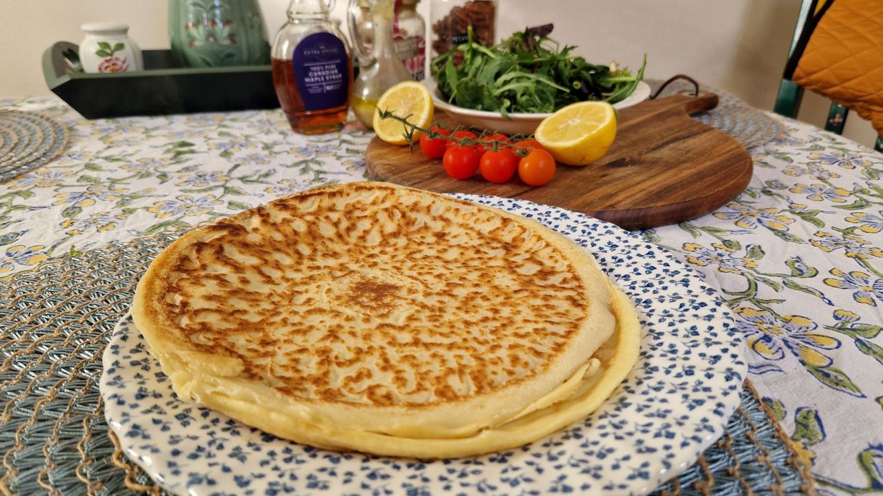 A plate full of untopped pancakes sits on a blue and white plate in the foreground. A chopping board with salad leaves, a sliced lemon and tomtaoes on the vine is in the background.