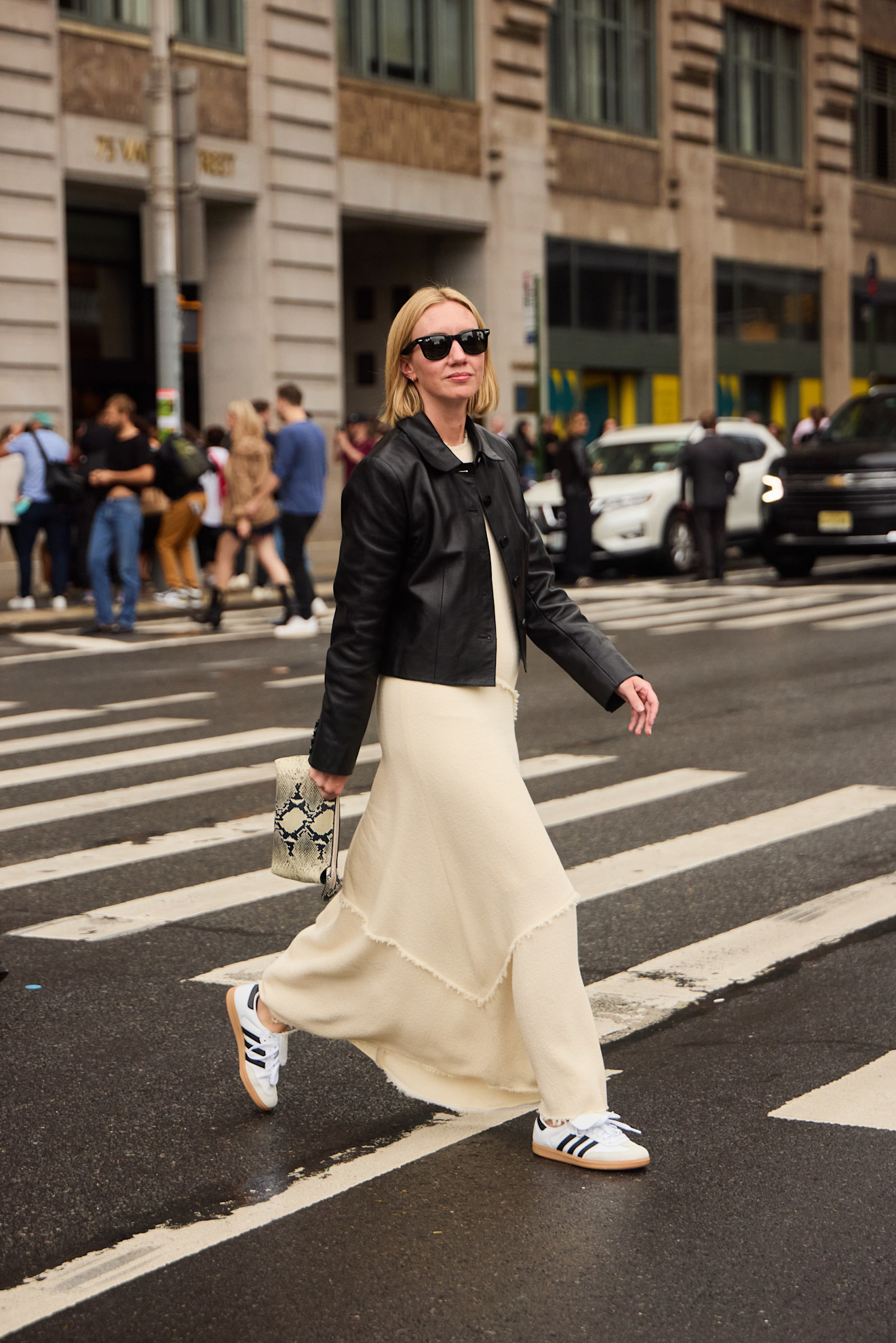a fashion week attendee crossing the street in sunglasses, leather jacket, off-white dress and adidas sneakers