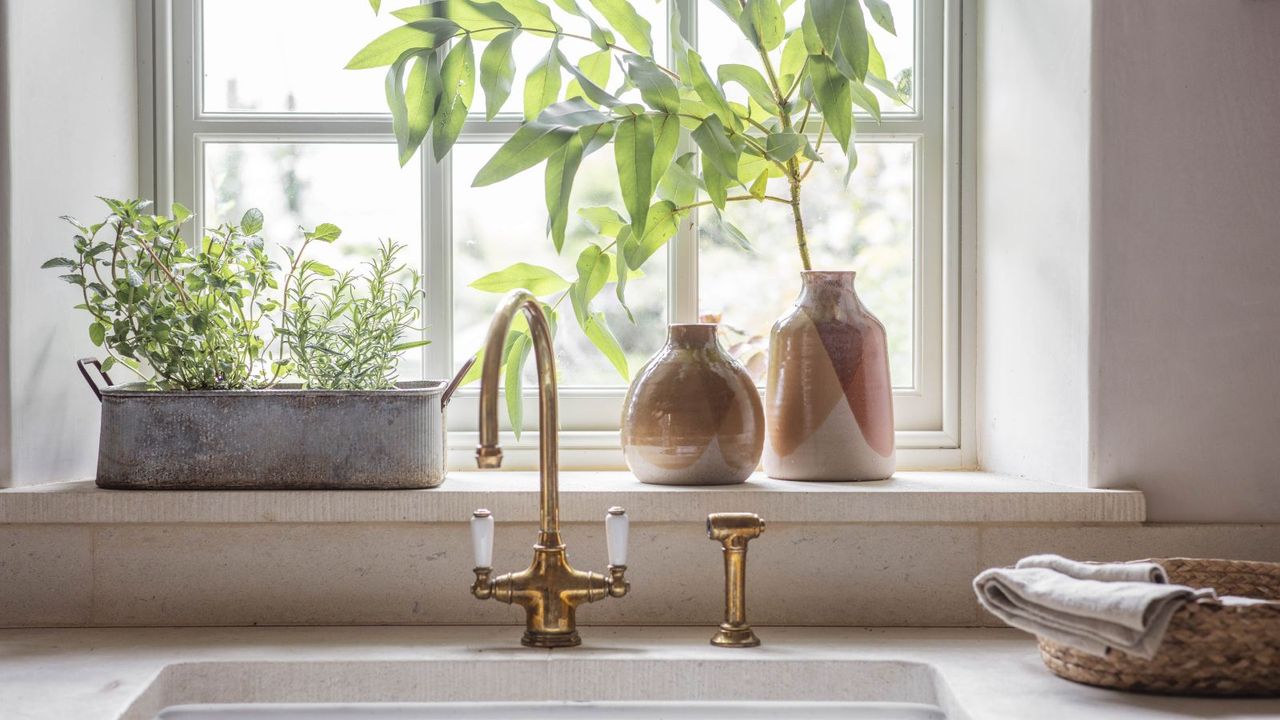 A kitchen sink inset into a stone countertop with a brass arched faucet. A window sill behind with some plants in earth ware vases and a metal trough. Some folded towels beside. 