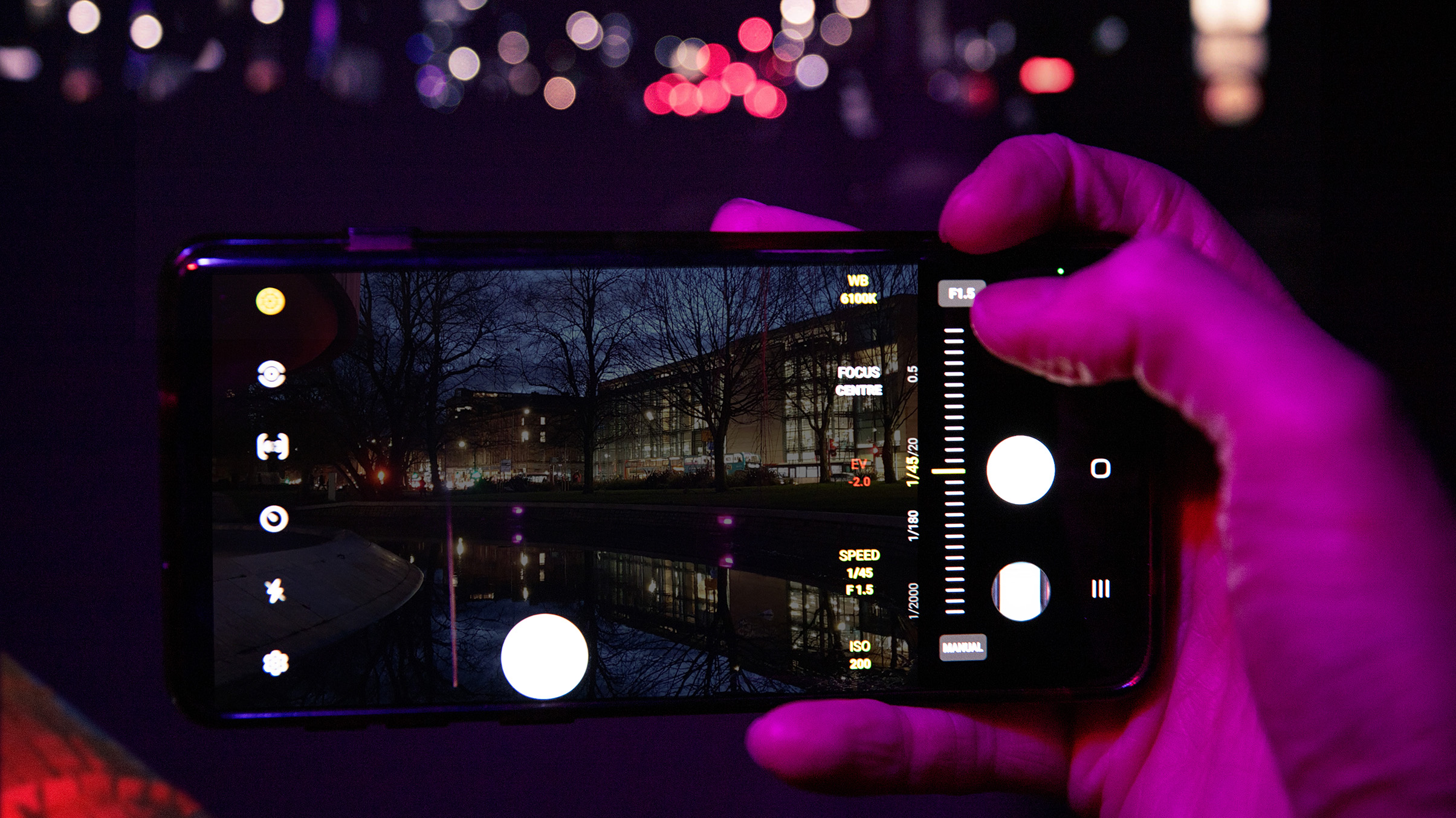 A hand holding a smartphone displaying a night-time camera interface, featuring settings for exposure, focus, and ISO