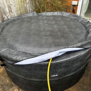 A grey faux-leather hot tub with lid being filled with a yellow hosepipe