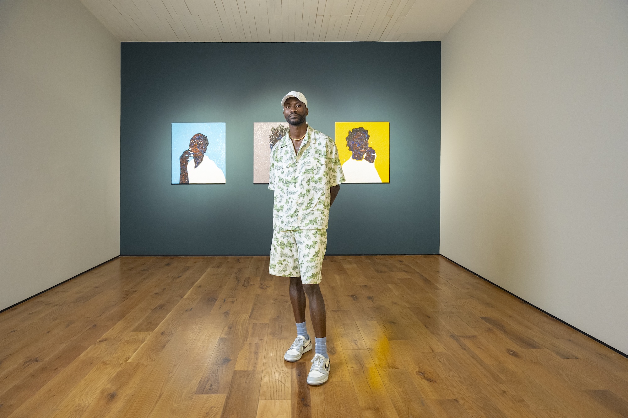A man wearing a light green floral short-sleeve shirt and matching shorts, a white cap, and sneakers stands in an art gallery with wooden floors and dark teal walls. Three colorful portrait paintings hang behind him, evenly spaced on the wall.