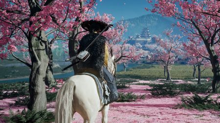 Ghost of Yotei gameplay showing Atsu sitting on her horse between bright pink cherry blossoms, looking at a distant fortification built against a mountain
