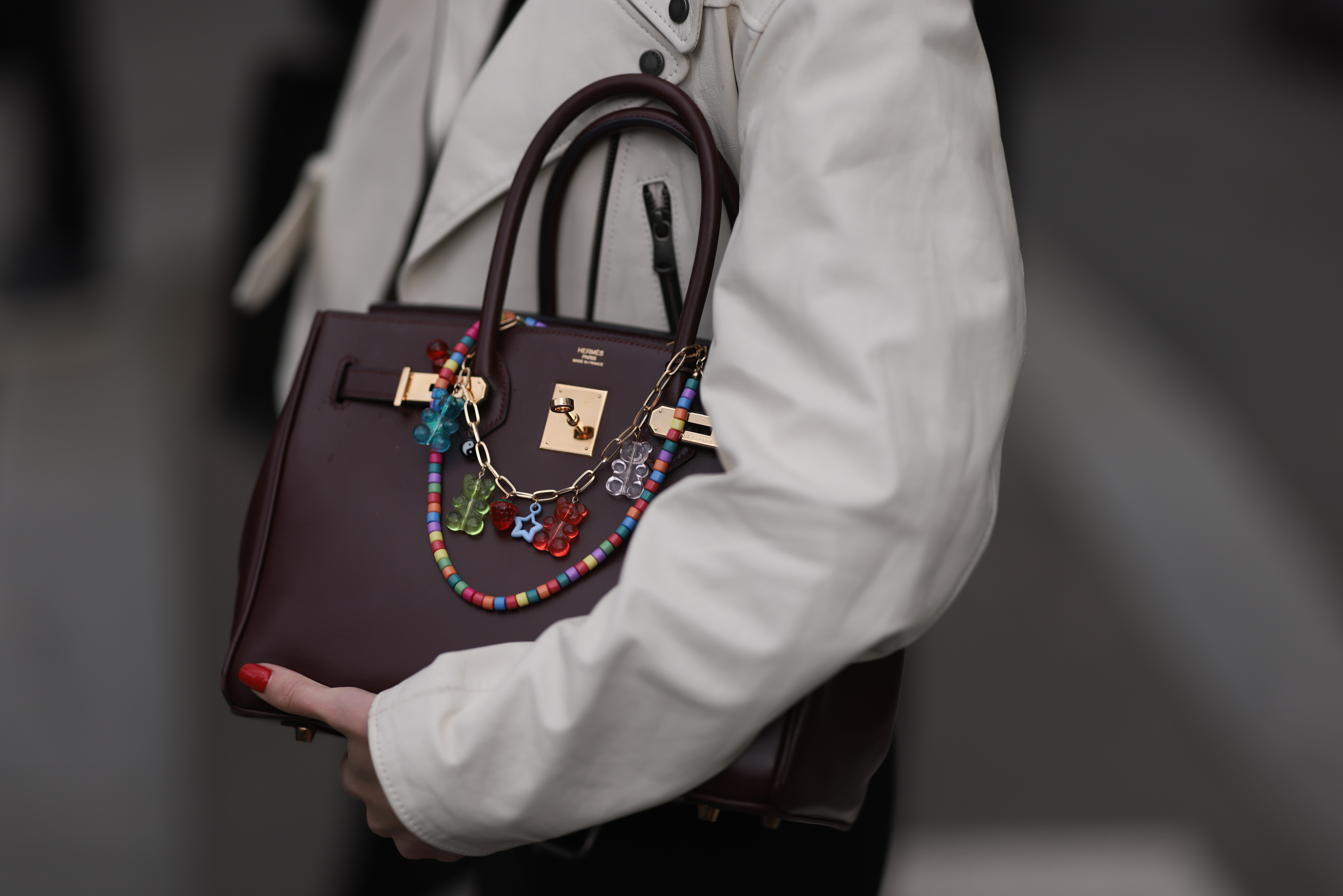 A guest seen wearing a white leather short trenchcoat jacket and a wine red Herm&egrave;s birkin bag with colourful bag chains outside Miu Miu Show