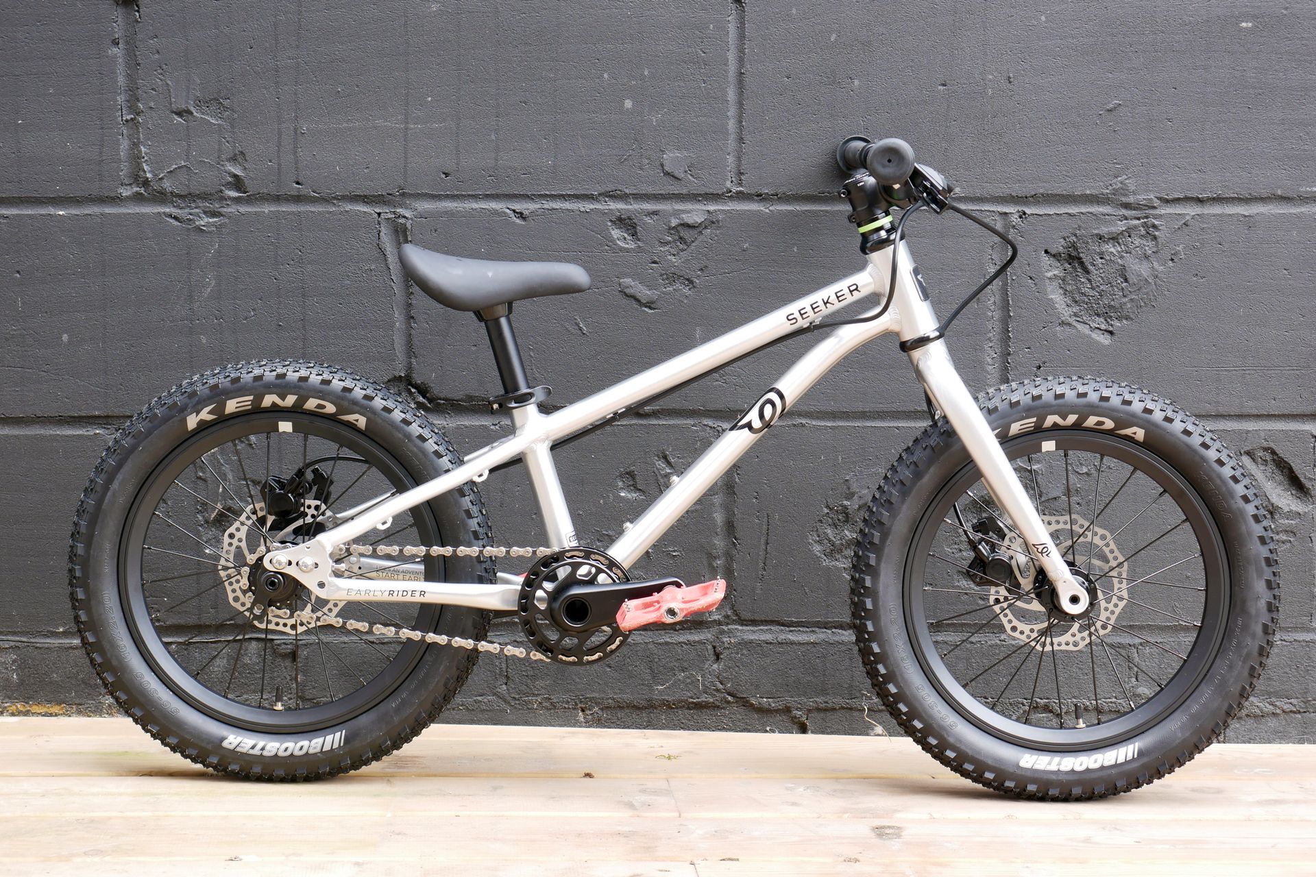 The Early Rider Seeker 16, with its polished silver finish, all black componentry, and Red Funn pedals, against the backdrop of a black painted breeze block wall