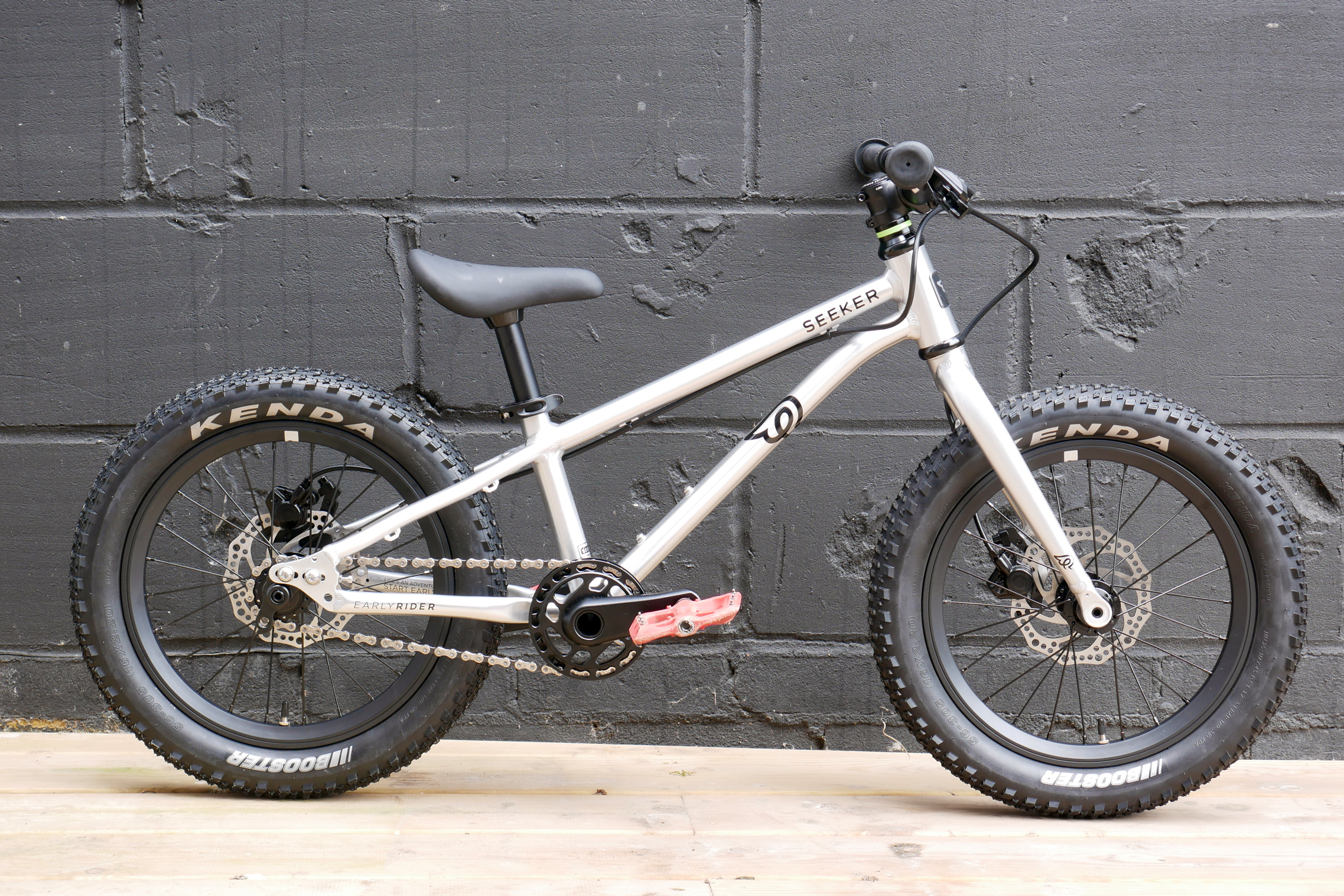 The Early Rider Seeker 16, with its polished silver finish, all black componentry, and Red Funn pedals, against the backdrop of a black painted breeze block wall