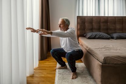 man performing a squat looking off camera, wearing a white sleeved top and blue trousers in a bedroom setting on a cream rug and wooden floors. there's a brown bed next to him and white long net curtains in front