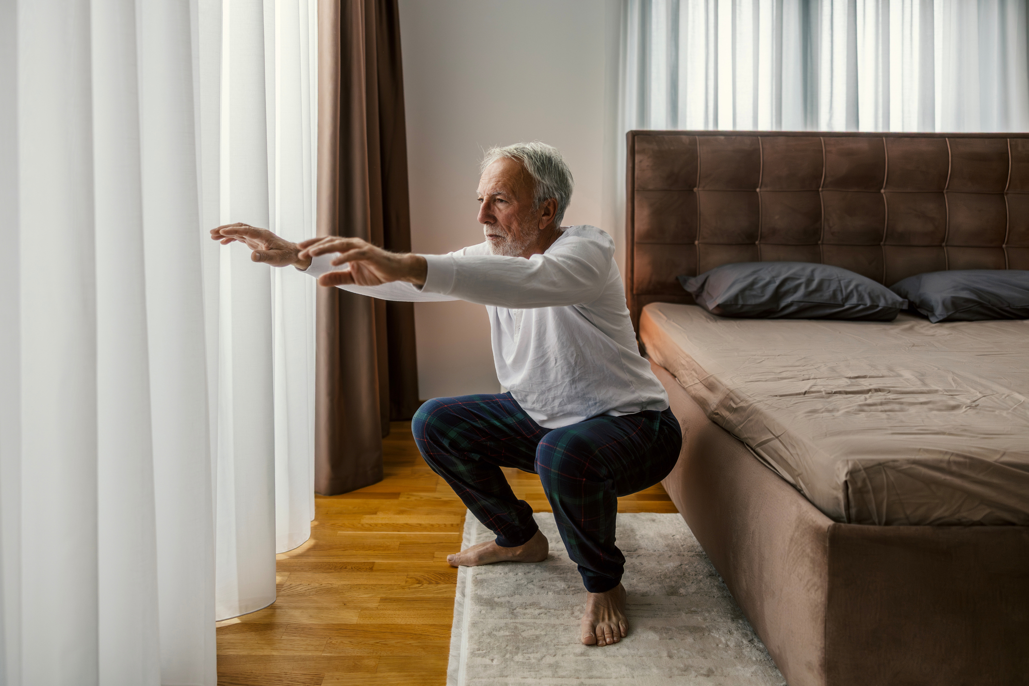 man performing a squat looking off camera, wearing a white sleeved top and blue trousers in a bedroom setting on a cream rug and wooden floors. there's a brown bed next to him and white long net curtains in front