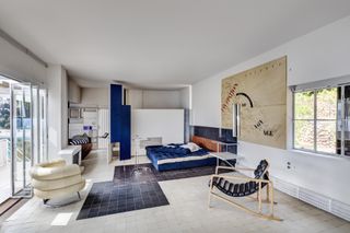 A bedroom with a blue double bed, a wooden headboard, a white leather chair, a lounger, a large map on the wall, a sliding door and black and white tile flooring.