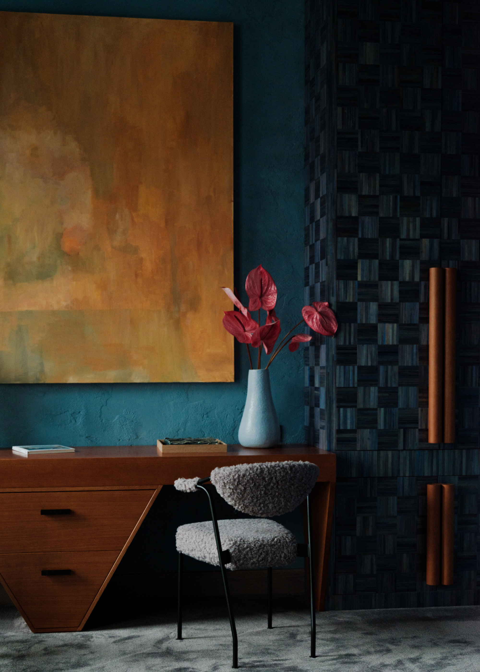 A blue study with a yellow painting, a blue checkerboard cabinet with brown handles, a wood desk, a fuzzy chair, and a vase of anthuriums