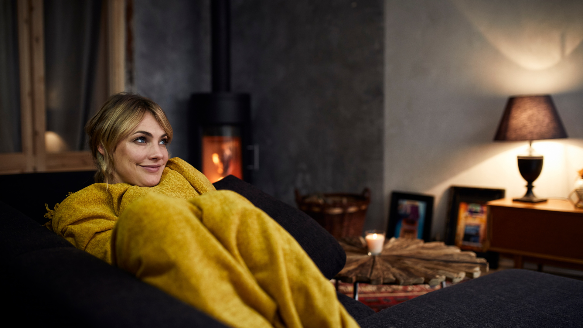 A woman sits on a couch in winter wrapped up in a yellow blanket.