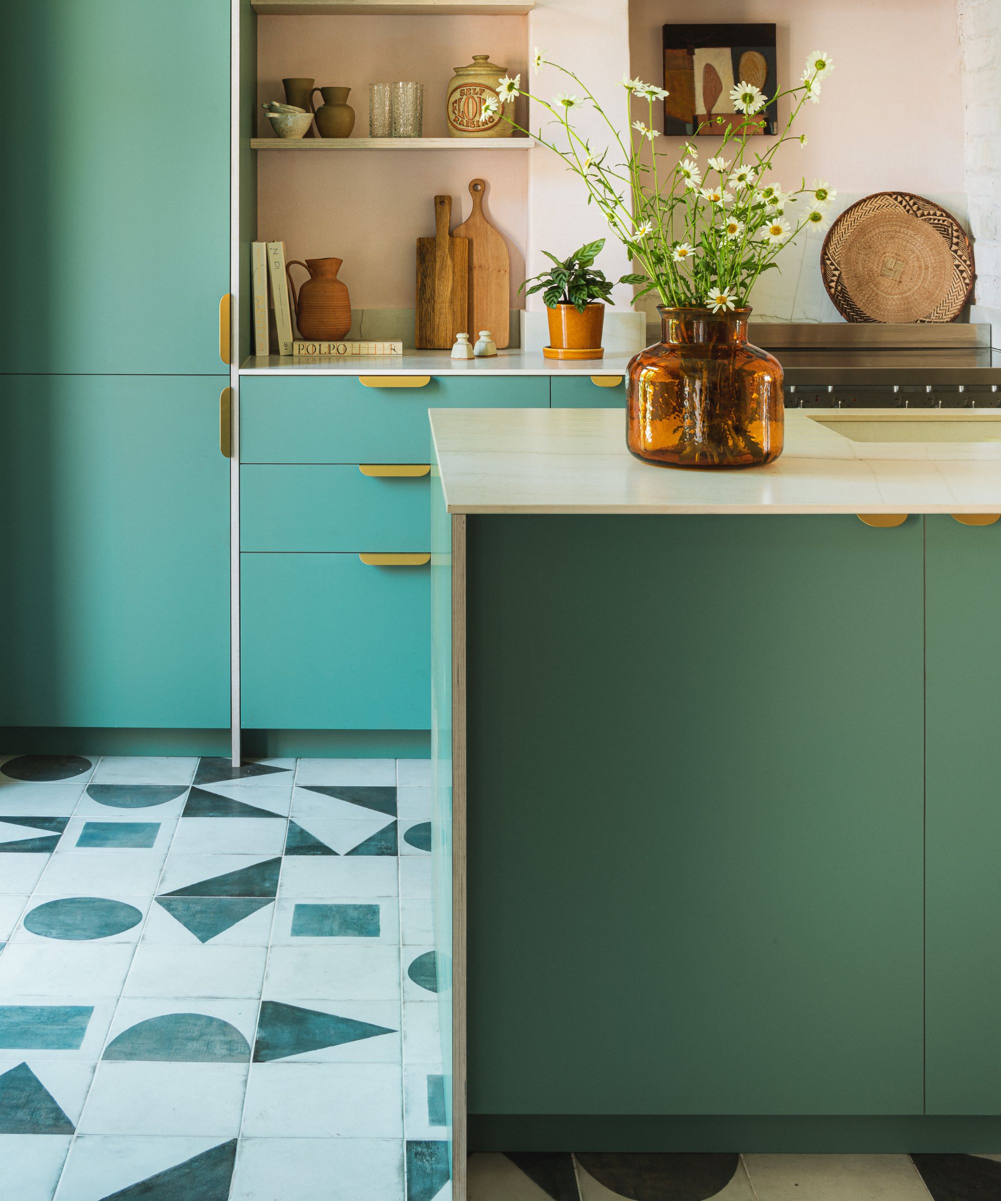 Playful modern kitchen featuring teal and forest-green cabinets, brass pulls, a pale worktop, and bold geometric patterned floor tiles.