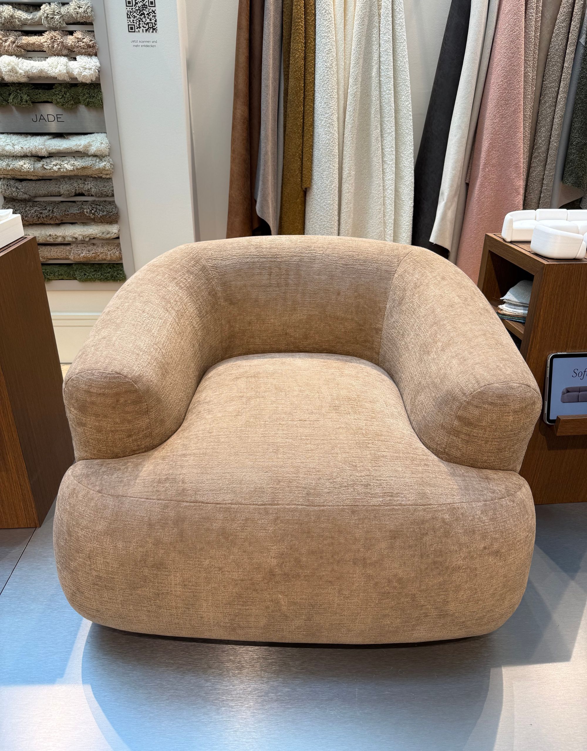 A round armchair in a beige colorway with hidden feet in front of a scarf-like display of sofa fabrics running along the wall behind