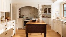A warm, traditional kitchen featuring a large arched alcove over the stove with a textured stone backsplash, off-white cabinetry with brass bin pulls, and a dark wood antique-style island in the center