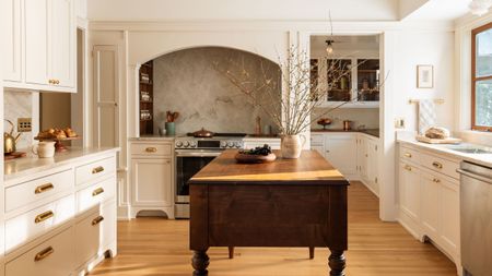A warm, traditional kitchen featuring a large arched alcove over the stove with a textured stone backsplash, off-white cabinetry with brass bin pulls, and a dark wood antique-style island in the center