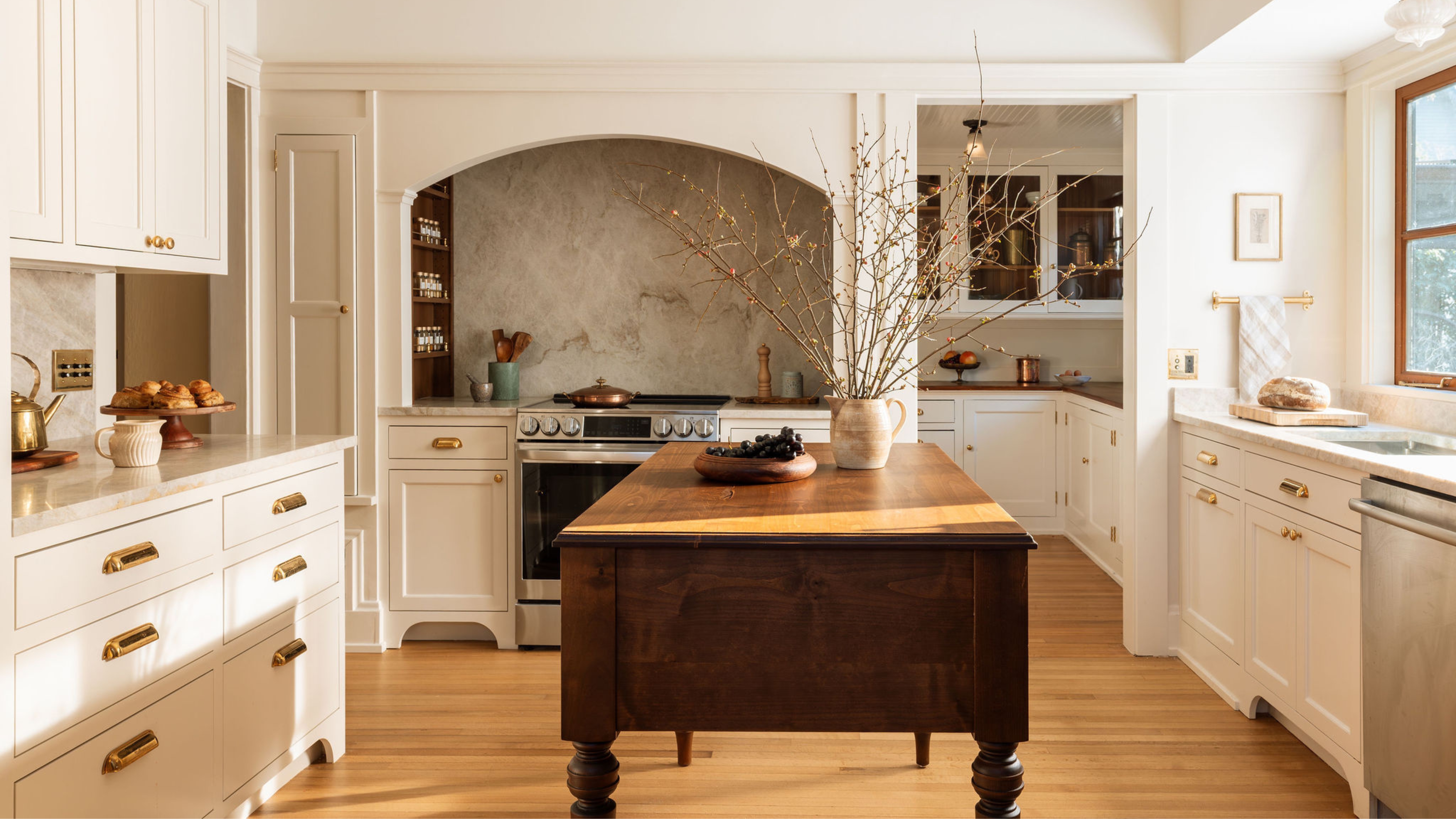 A warm, traditional kitchen featuring a large arched alcove over the stove with a textured stone backsplash, off-white cabinetry with brass bin pulls, and a dark wood antique-style island in the center