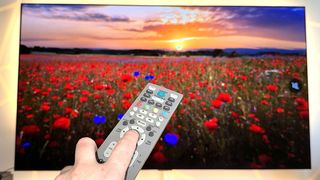 An outstretched hand holding a remote control in front of a 65-inch TV, which is mounted on the wall