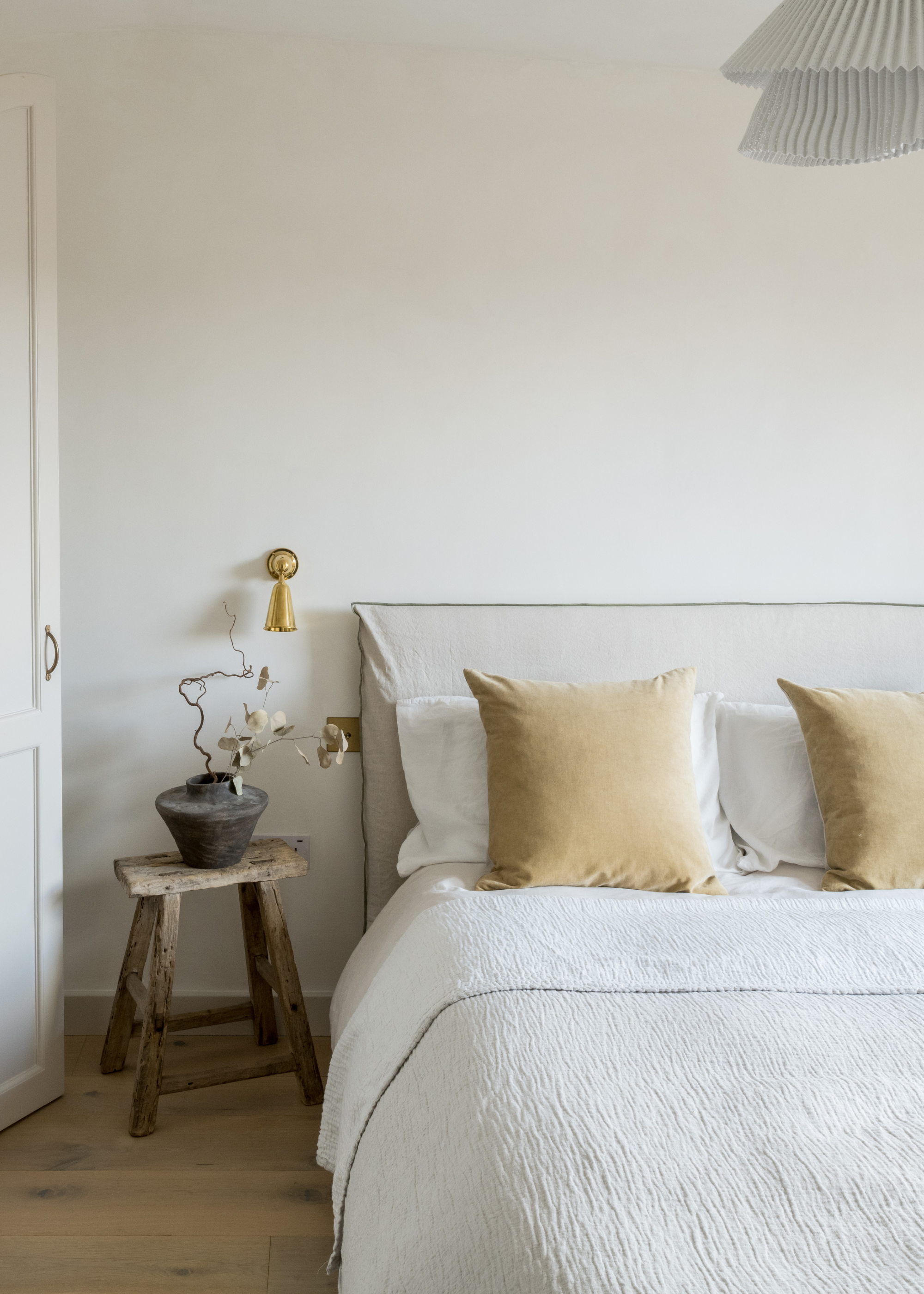 A bright bedroom with a pleated pendant light, a white bedscape, yellow cushions, white pillows, gold wall sconces, a wood stool, and a vase with stems