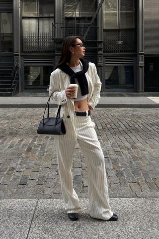 How to wear a suit is shown in a photo of a woman standing on the sidewalk in New York, wearing black sunglasses, silver earrings, a white and black pinstripe blazer over a white cropped tank top styled with matching pinstripe trousers, a black belt, black tabi flats, a black shoulder bag, and a black cardigan layered over the shoulders