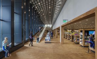 Interior view in the Eemcentrum building, wooden floor, visitors, glass framed wall to the left, stairwell, book cases and books, ceiling spotlights