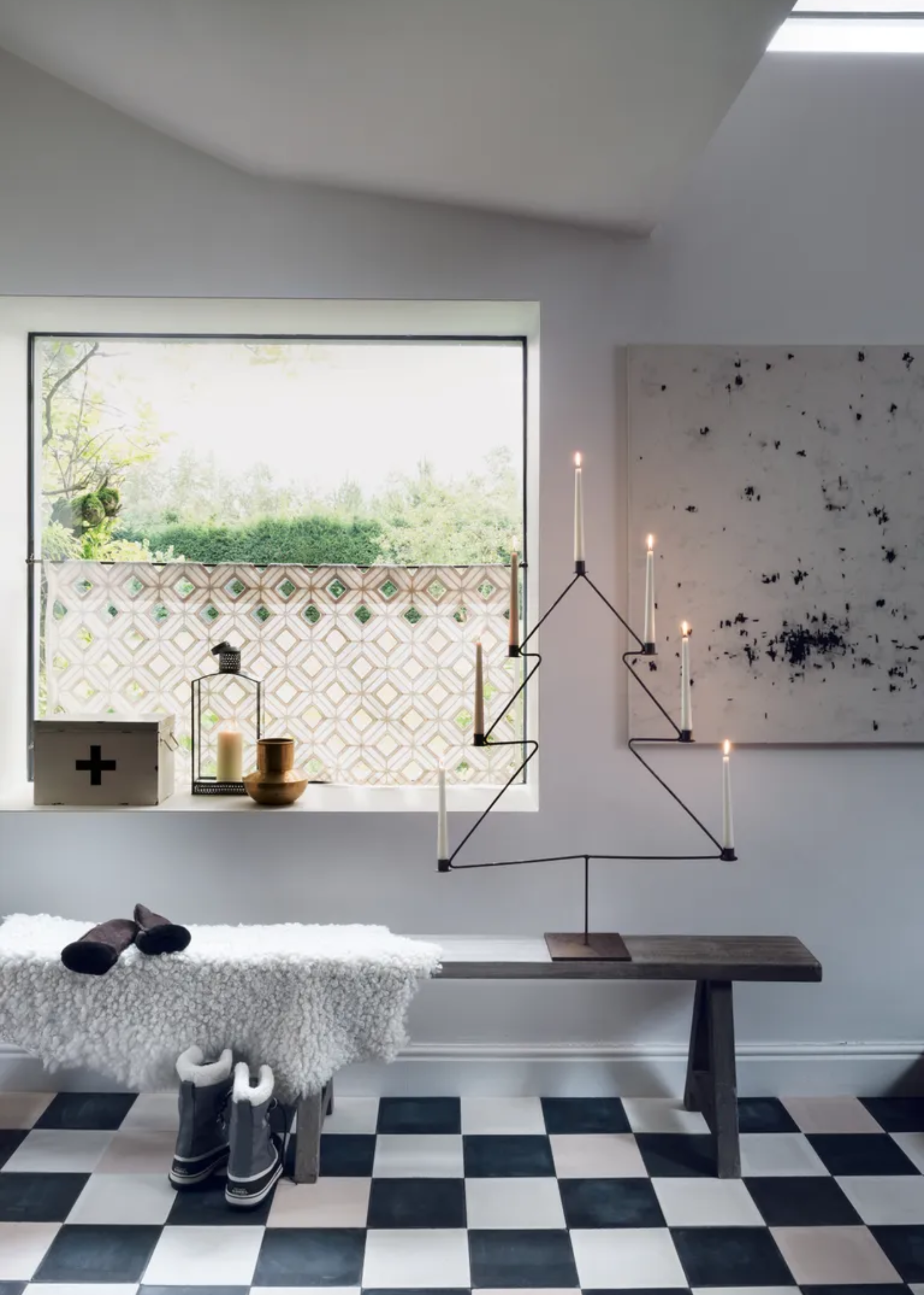 A white entryway with checkerboard floor tiles with a metal christmas tree candelabra, boots, a bench and a faux fur throw