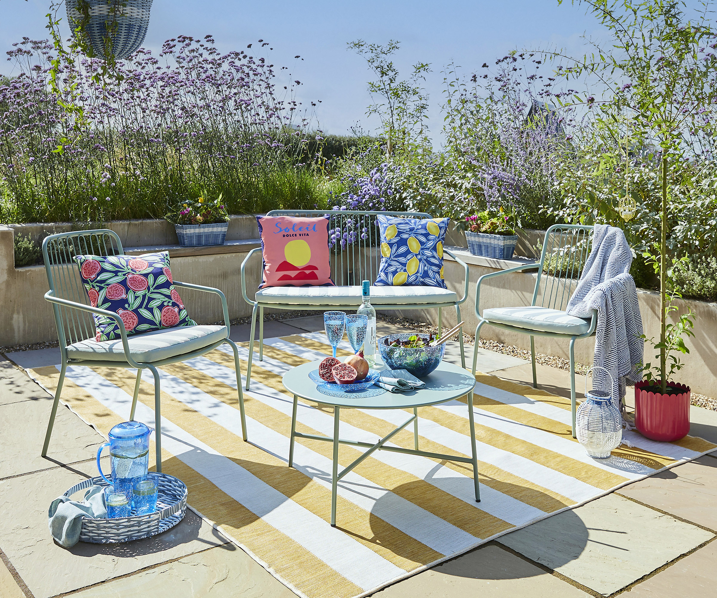 A sunny terrace with metal garden furniture with quirky outdoor cushion and throws in pastel shades