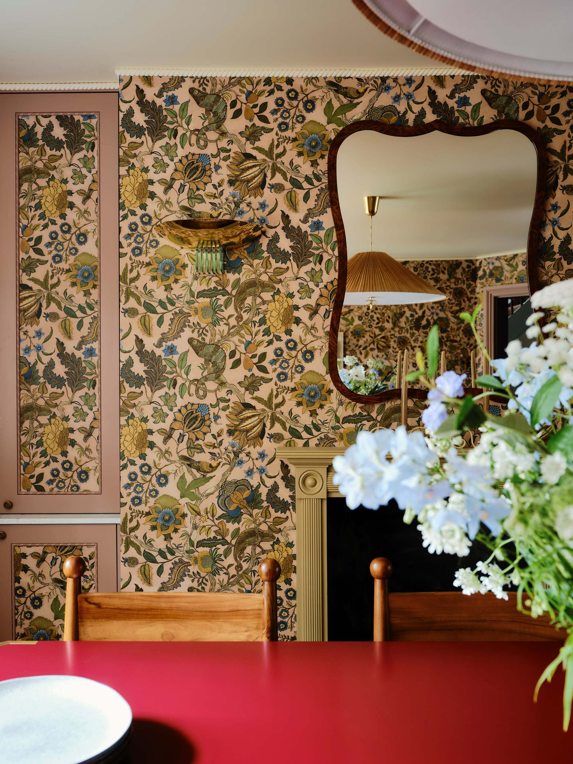 Image of a pink, green, and blue floral wallpaper in a dining room with a mirror over the fireplace and a red dining table.