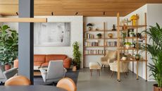 an open plan living room with timber clad ceiling, white brick walls, open timber shelving that extends to a room divider, polished concrete floors, shearling armchairs, a tan leather sofa and gray armchair, and a black dining table with tan dining chairs